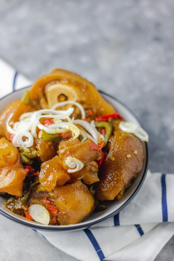  cow leg (cow foot/bokoto)in a black bowl. peppered cowleg garnised with sliced onions. the bowl is placed on a napkin.