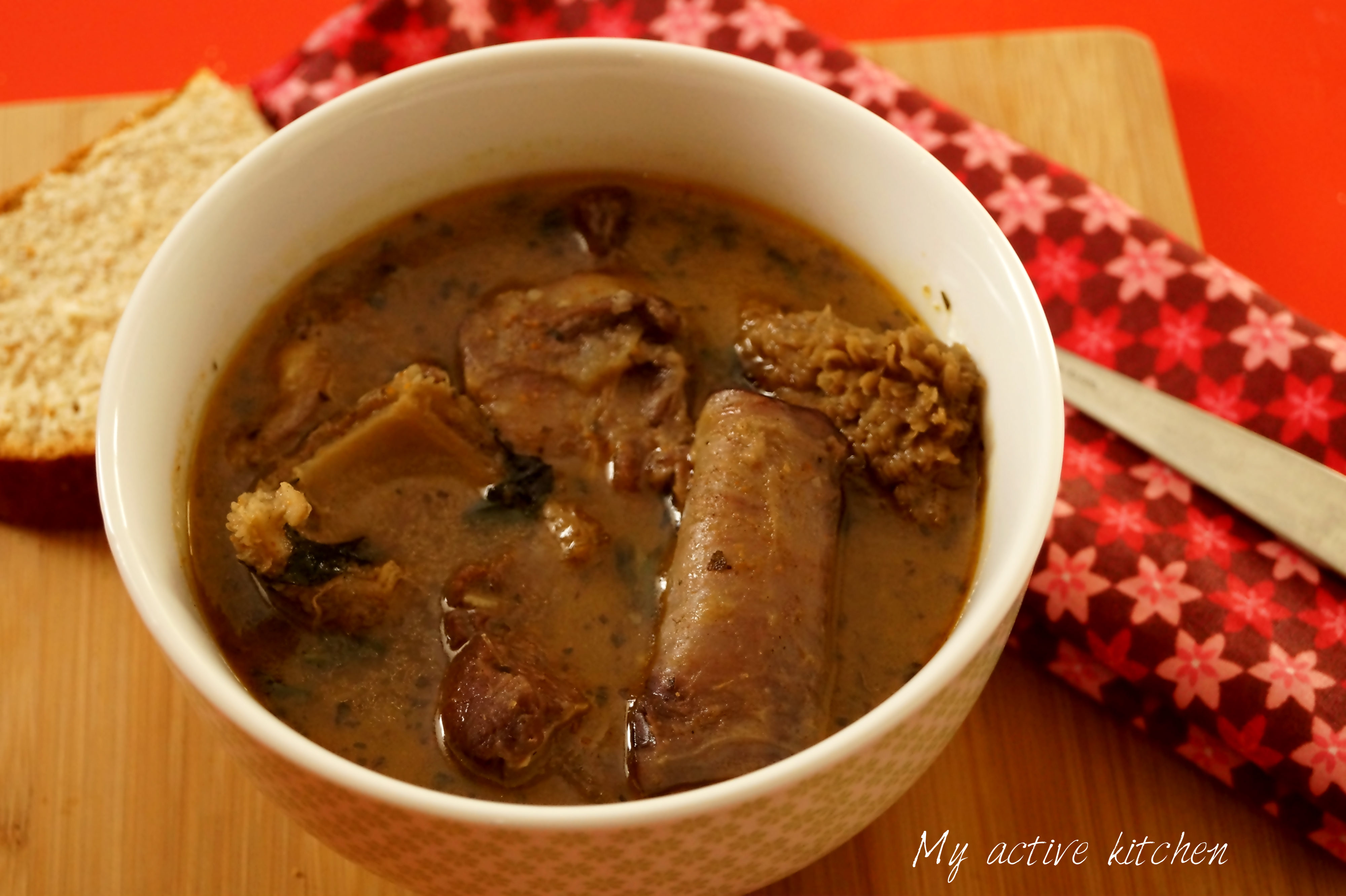 Nigerian assorted meat pepper soup in a white bowl placed on a red table. The bowl is placed beside a floral napkin and crusty bread