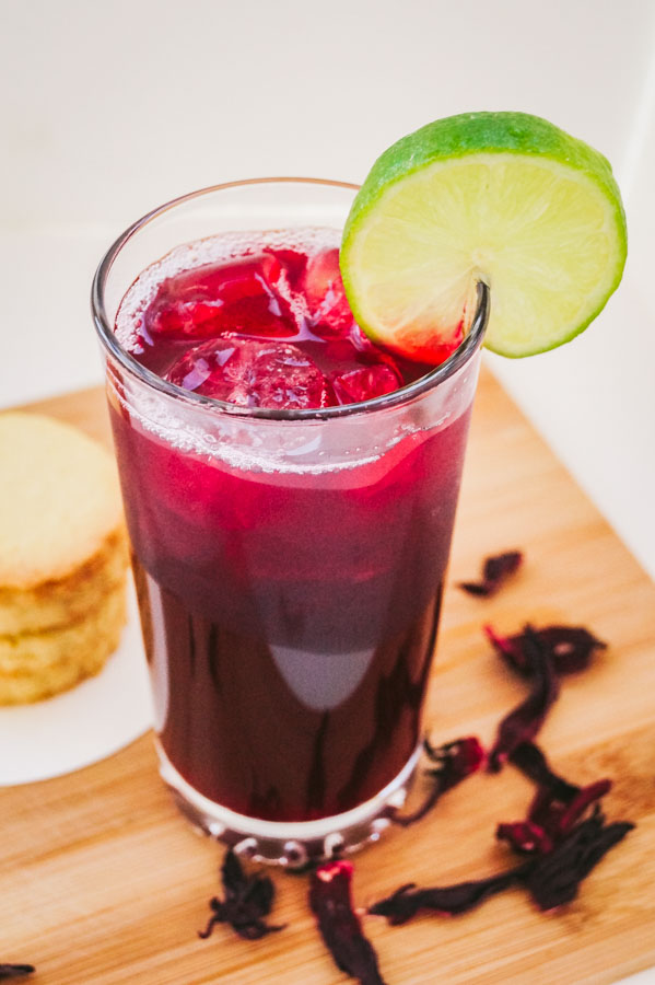 an angled overhead shot of zobo drink garnished with a slice of lime, beside it are stack of cookies