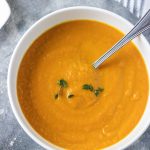 Thick and creamy healthy carrot leek soup served in a white bowl and garnished with fresh thyme. Ready to be eaten with a crusty bread roll on the side