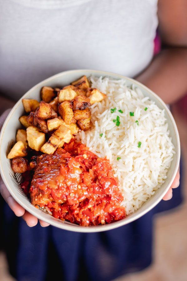hand holding a bowl of basmati rice served with fried stew and fried plantain.