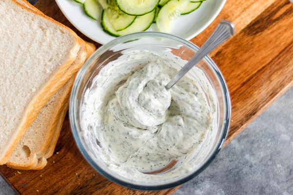 cucumber sandwich filling in a bowl with slices of soft white bread and cucumber slices on a board.