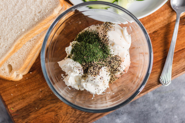 cream cheese, dill, black pepper and mayonnaise in a bowl placed on a board alongside slices of bed.